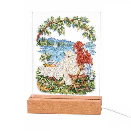 Lakeside Lounge Under a Leafy Arch light-up pearl painting – Red-haired woman lounging by a blue lake under a leafy flowered arch, beside a picnic table