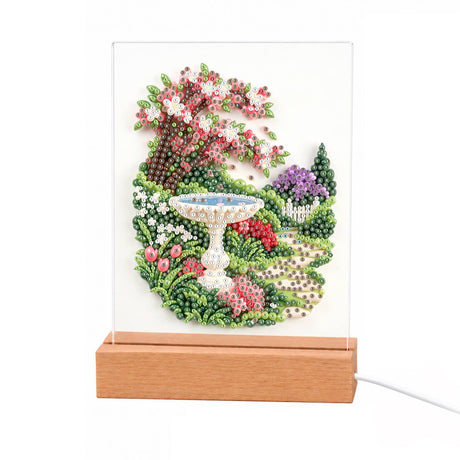 Stone Birdbath Under Pink Blossoms light-up pearl painting – White birdbath on a garden path under a pink blossom tree, with a picket fence and flowers