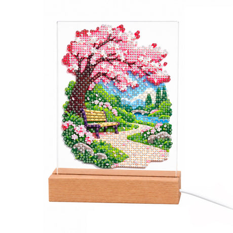 Cherry Blossom Path to the River light-up pearl painting – Pink cherry blossom tree arching over a park bench and stone path by a river and mountains